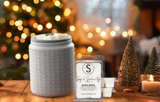 Soy & Serenity wax melts displayed beside a gray wax warmer on a wooden table, with soft golden Christmas lights and miniature holiday trees glowing in the background.