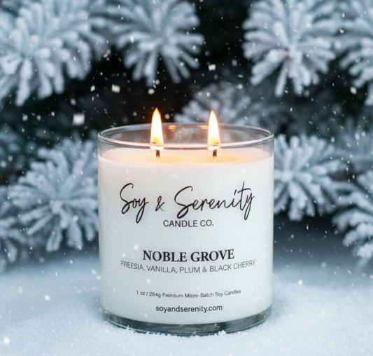 Soy & Serenity Noble Grove soy candle with vanilla, plum, and black currant, burning in front of snowy evergreen branches; clean, non-toxic winter candle.
